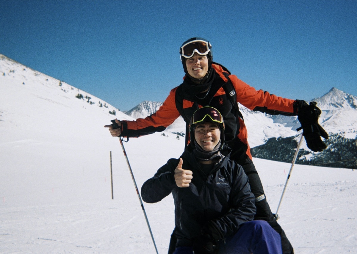 Two friends skiing on a mountain, one giving a thumbs up with snowy peaks behind