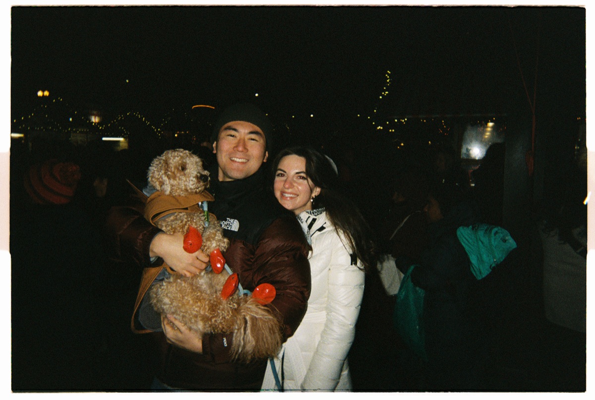 Couple holding a small dog at a nighttime outdoor event, city lights in the background