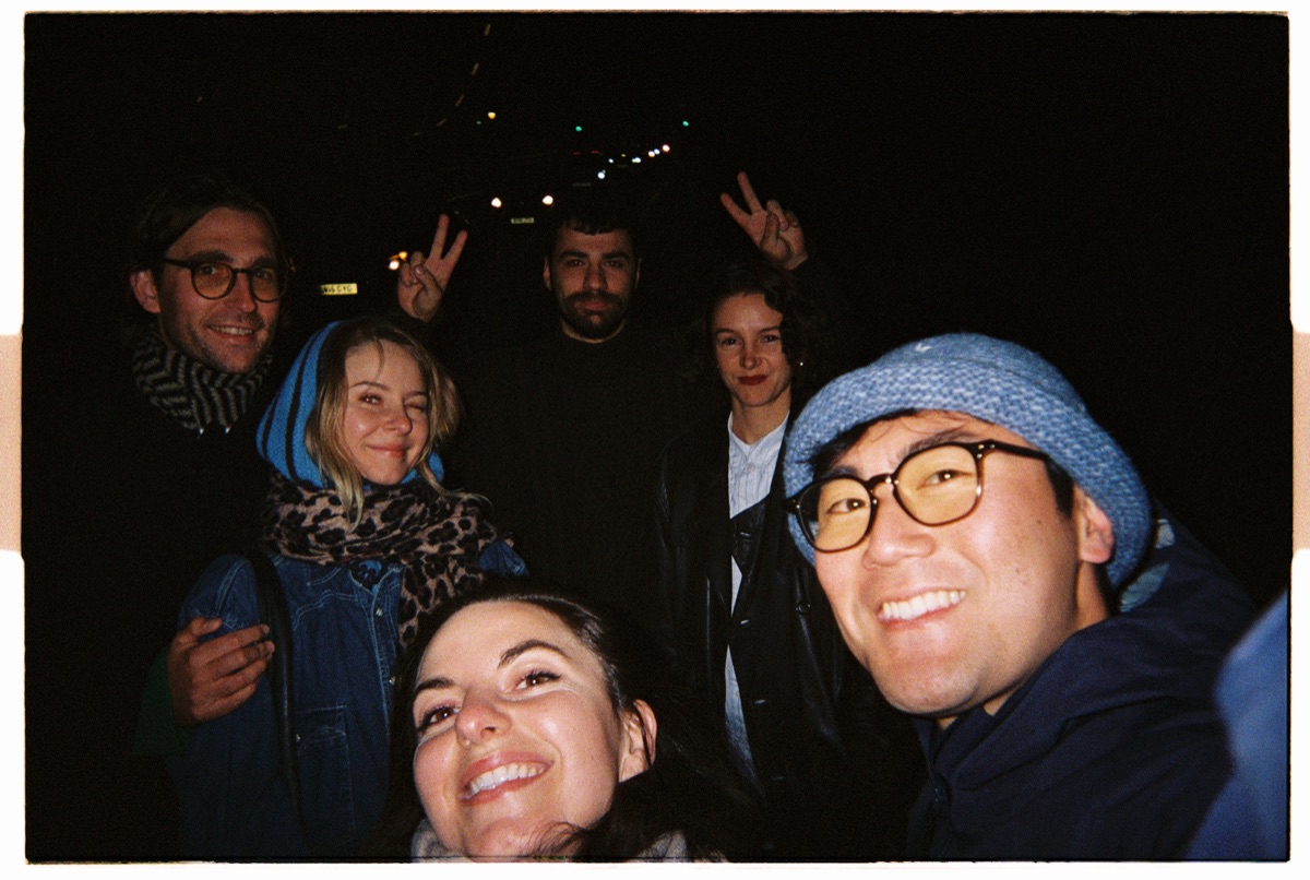 Group selfie of six friends outdoors at night with string lights behind