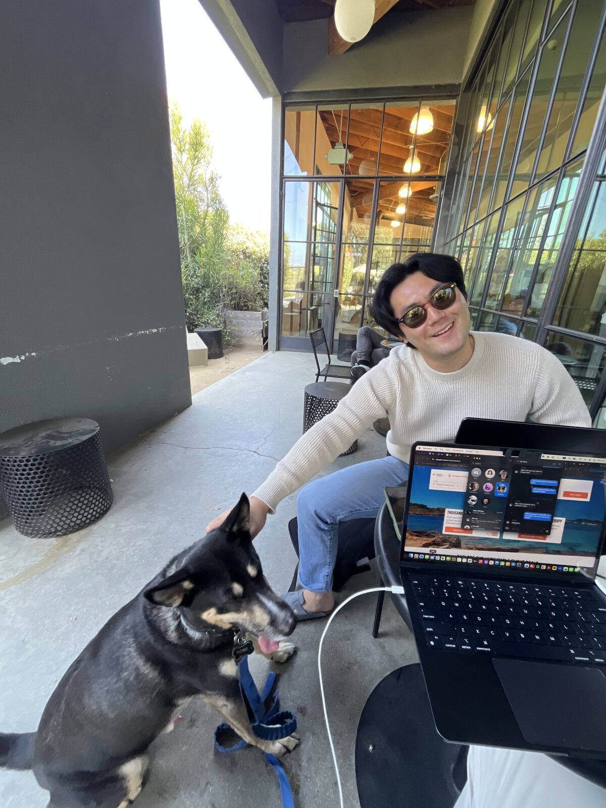 Kevin at an outdoor cafe table with a laptop open to a video-call grid, Kobe the dog on a blue leash leaning against his leg