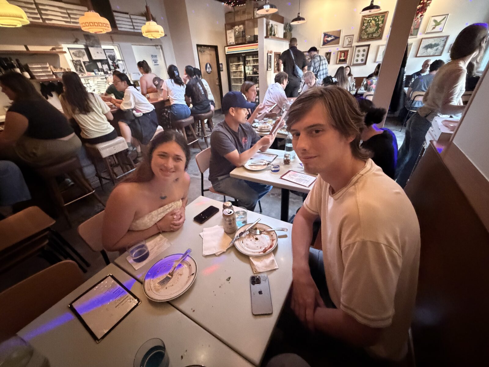 Dakota and Maddie smiling at a table in a dim, busy restaurant, plates and phones in front of them
