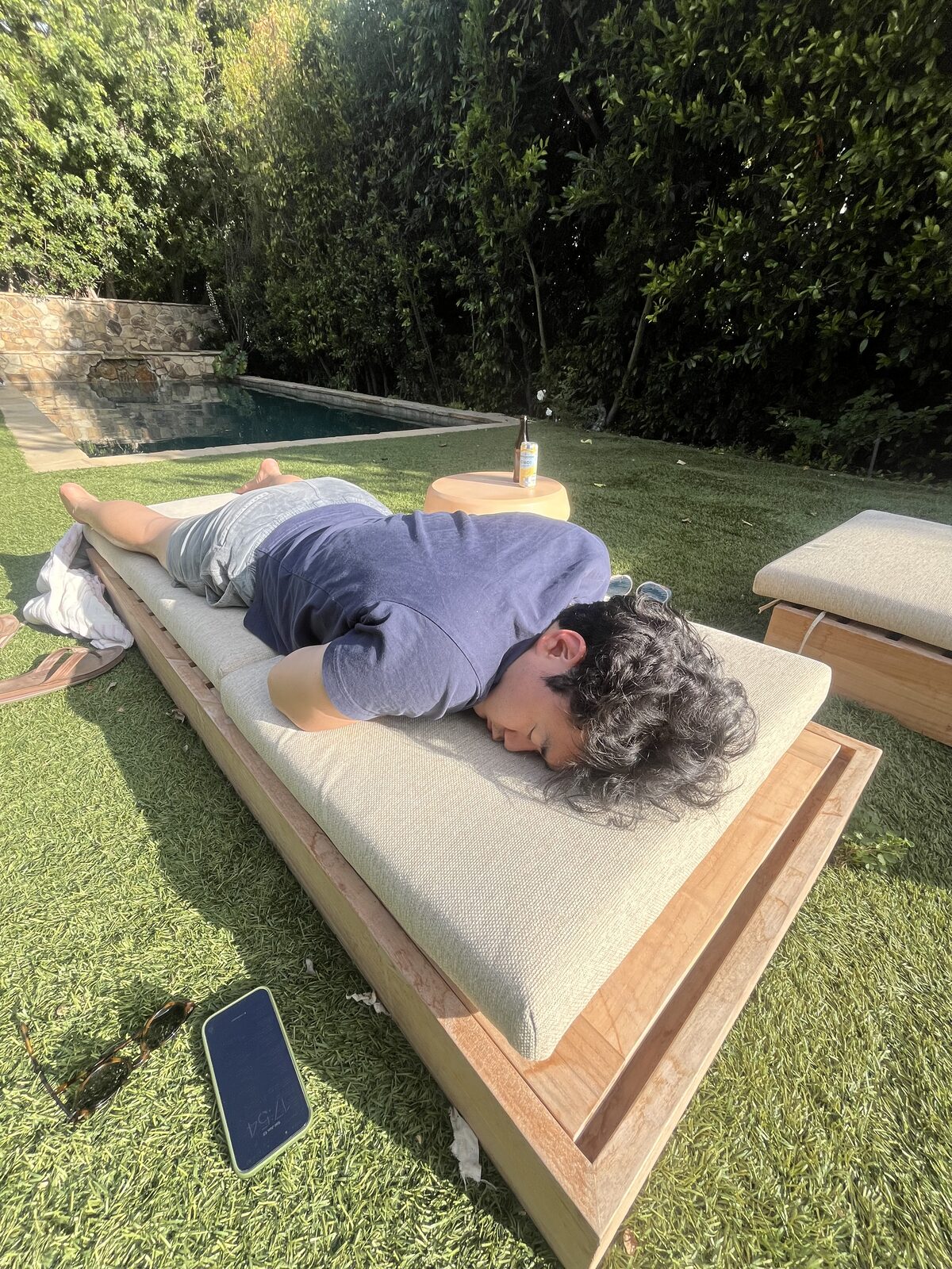 Kevin asleep face-down on a wooden outdoor daybed in a sunny grassy backyard, phone lying on the grass nearby