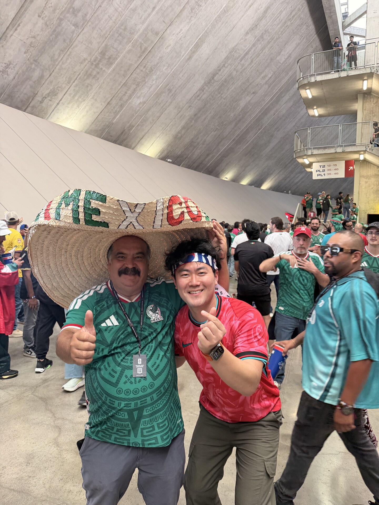 Kevin in a red Korea jersey throwing a Korean finger heart next to a Mexico fan in a giant MEXICO sombrero and green jersey in the Estadio Akron concourse