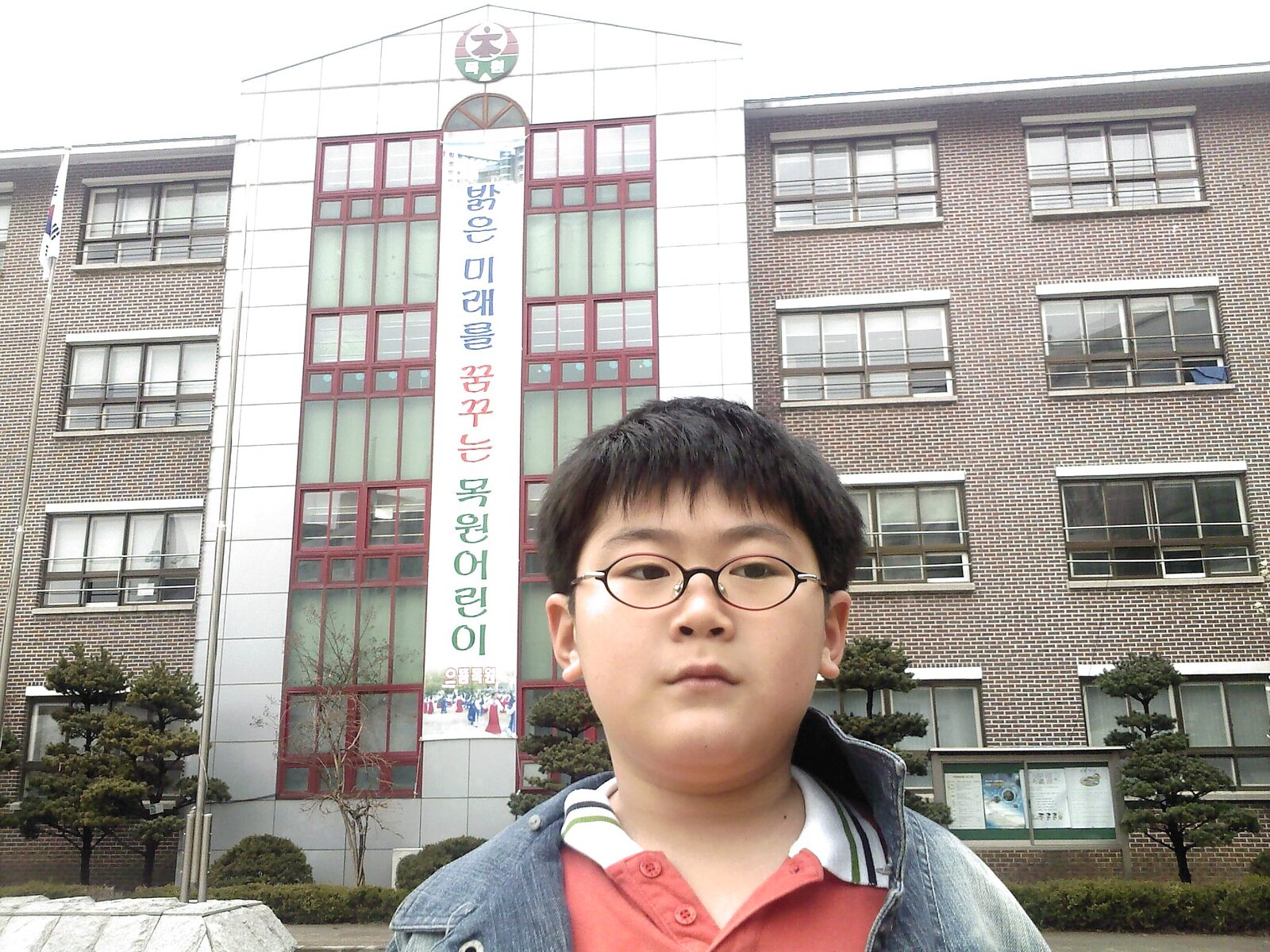 The original photo: young Kevin in oval glasses and a denim jacket in front of Mokwon Elementary School in Korea